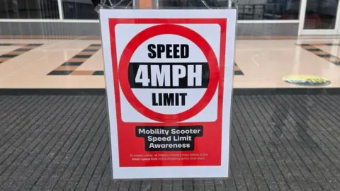 A sign in the window of Idlewells Shopping Centre which shows 4mph, and says "mobility scooter speed limit awareness"