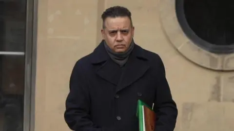 Shahid Malik pictured leaving court. He is wearing a navy overcoat and carrying some files.