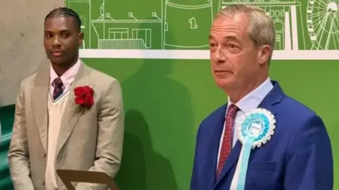 Martin Suker Jovan Owusu-Nepaul, in a cream coloured suit with a shirt and tie on, standing next to - and looking at - Nigel Farage, who is wearing a navy coloured suit with a shirt and tie on.