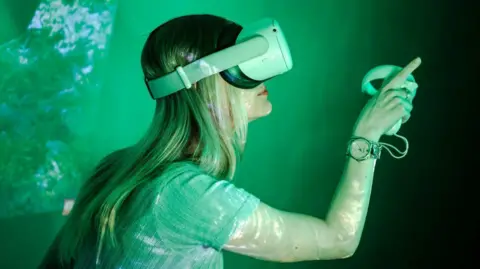 ARU A woman with long, fair hair is wearing virtual reality goggles and holding a controller in her right hand. She is wearing a short-sleeved top and is pictured against a green backdrop.