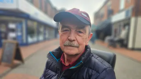 Steve is wearing a burgundy cap and a navy padded coat. He has a moustache and is sitting in a mobility scooter. 