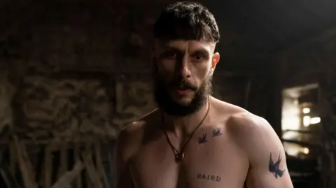 BBC/Mam Tor Productions/Anne Binckebanck Richard Gadd in character as Ruben Pallister. He is shirtless, with tattoos on his arm, chest and shoulder blade. He is wearing a gold necklace. Planks of wood lean against the exposed brick of a barn, as sunlight leaks through the gaps in a boarded-up window.
