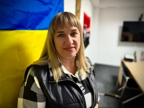 Ira Savka is wearing a black white and grey checked shirt with a black gilet as she looks up to the camera in an office. A desk and chair is behind her and a Ukrainian flag is hanging up.