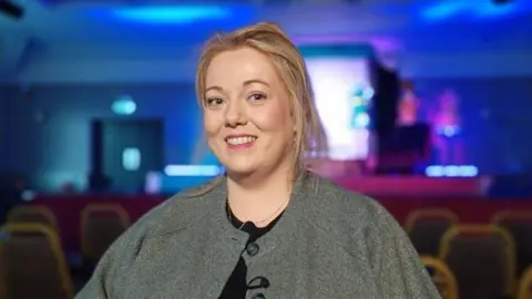 Nuala Moore, a woman with fair hair tied back, wearing a grey shawl-style jacket and black top.