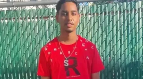 Family  Shawn Seesahai in a red tshirt with a large letter R on it stood in front of a green fence