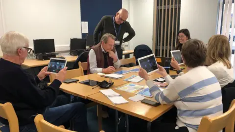 Molly Pipe/BBC Five people are sat around a table and one man is standing near the table. Four of the people are looking at digital tablets on which there is a video call. In the background there is a row of three desktop computers.