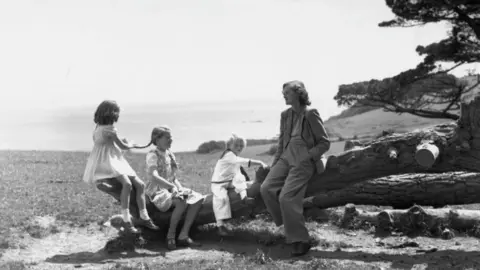 J Wilds/Keystone Features/Getty Images A black and white photo showing Daphne du Maurier sitting on a tree trunk with a young boy in a sailor suit and two young girls wearing dresses. Du Maurier is wearing a trouser suit. One of the girls is holding one of the other's plait. Behind them is a cliff and the sea beyond.