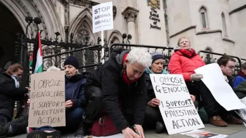 PA Media Several people sit outside the Royal Courts of Justice holding signs reading "I oppose genocide, I support Palestine Action"
