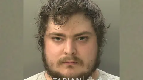 A mugshot of a man with lots of brown hair and facial hair. he is looking intensely, directly into the camera.