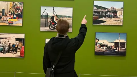 Tom Hope PR Ian Watkins at The Valleys exhibition. He is looking at a wall of colourful photographs depicting valleys life in the '90s. He wears a black coat, black and cream scarf, black glasses and has blond short hair. The photograps are from Tina Carr and Annemarie Schöne from their 1991 series Coalfaces: Life After Coal in the Afan Valley