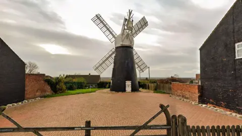 A black brick windmill with white wooden sales stands on a red-brick paved area. There are buildings on both sides and a fence in the foreground.