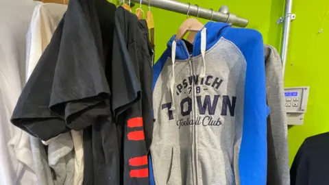 Laura Foster/BBC A blue and grey Ipswich Town Football Club hoody hanging on a silver rail alongside dark t-shirts