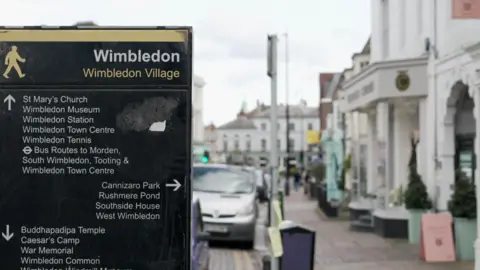 A street sign in Wimbledon Village with directions to places of interest. There are parked cars and buildings in the background.