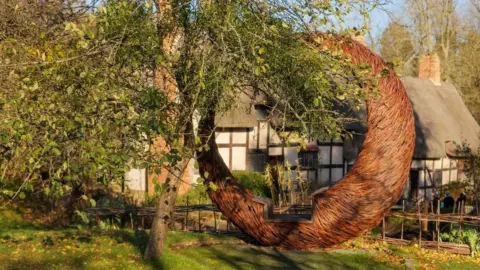 Shakespeare Birthplace Trust A large circular seat shaped like a hollowed out full moon is in a garden with the house in the background. It is brown and looks to be made of weaved wood