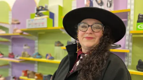 Emma Baugh/BBC A woman wearing glasses and a large black hat in front of a colourful display of children's shoes.