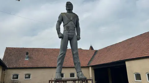 Franz Josef Zika A worn-looking Michael Jackson statue situated in a courtyard. Parts of the statue look brown and stained white. It is supported by nearby safety cables
