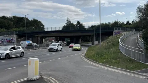 Leeds City Council Cars go by on a three-lane road. A traffic bollard is in the foreground. To the side is a footpath which slowly escalates uphill.