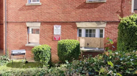 A brick-built house with its lower floor windows boarded up. The house is framed by green bushes in a garden area.