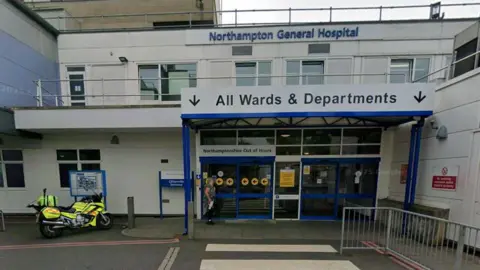 Entrance to hospital, a two-storey white building with white-framed windows. There are four blue doors, with a white one in the centre. A white sign with dark blue writing indicated "All Wards and Departments". There is a grey railing to the right and a motorcycle with rider in yellow hi-vis to the left.