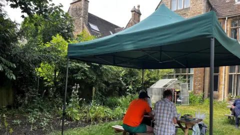 Emma Baugh/BBC A green gazebo with two people sitting underneath it, in a garden, in the background is a stone building.