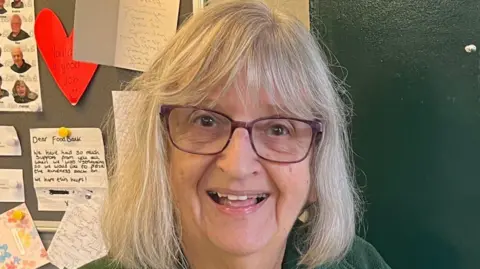 Jane Calcott, una mujer parada frente a un cartel, sonríe. Tiene el pelo hasta los hombros y usa gafas. Detrás de ella había una nota pegada en el pizarrón. 