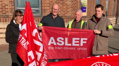 Four people stand on a street corner holding red banners and flags bearing the words: Aslef the train drivers' union. The three men and a woman are smiling and standing in front of a large red brick building.