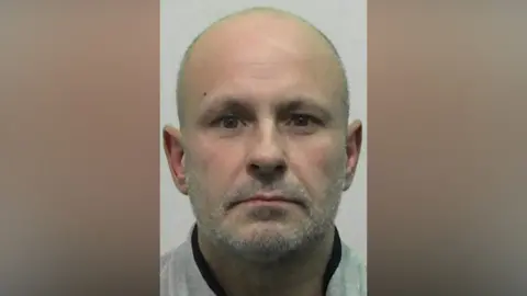 Mugshot of Adams. He is bald with a grey stubbly beard.