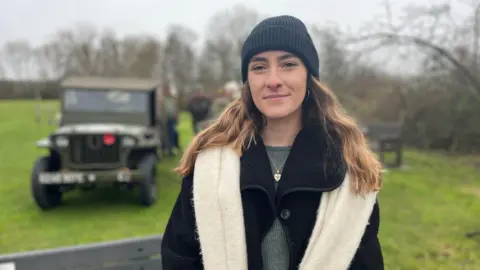 Camille Dahmen, a woman with wavy blonde hair, wearing a black hat and coat and a white scarf, looking straight at the camera. There's a stretch of grassland and a green wartime vehicle behind her.