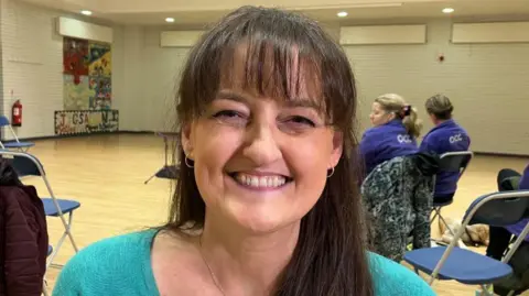 Beverley is smiling at the camera. She has long brown hair and has a fringe. SHe is wearing a bright blue top.