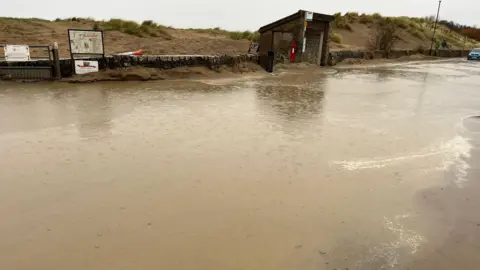 Jacqui Pool Flooding on Marine Parade in Instow on a rainy day. Sand dunes are in the background along with a bus stop shelter and red post box.