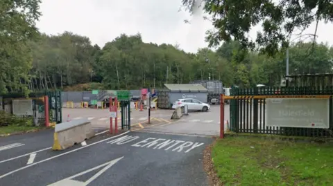 Google A road including the words no entry in white on the ground. Just beyond those words is a fence and a sign that includes the words welcome to Halesfield household recyling centre. Many trees are in the background.