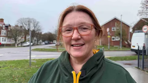 A woman with red hair and orange horn rimmed glasses smiles at the camera. She's wearing a Green and Yellow "Scouts" hoodie
