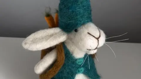 Mandy Smith/needlefeltingnook A needle felted white rabbit with a tall teal hat and carrots peeping out of its rucksack.