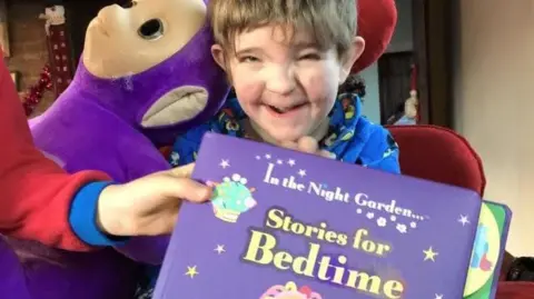 PA Media Jack Moate is grinning at the camera and is sitting in a chair with a high back and head support. He is wearing a bright blue top and has an Iggle Piggle purple toy on his shoulder while someone holds an In The Night Garden Bedtime Stories book in front of him.