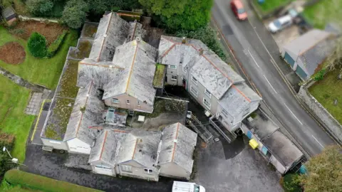 An aerial view of Fowey Community Hospital. It is a collection of grey buildings with a larger one in the centre and smaller ones surrounding it. There is a road on one side and garden on the other. 
