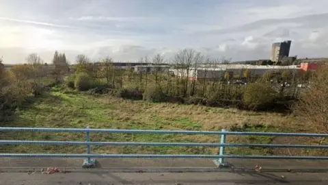 A view from a bridge of a parcel of unused land bordered by trees and hedges. A large B&Q store can be seen behind the trees.