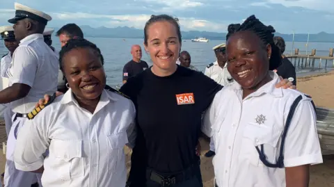 London Fire Brigade A female firefighter, wearing a black uniform, is standing in the middle of two female Malawi Police Force delegates, who are wearing a white uniform. They are stood on a beach in front of the sea with other delegates in the background. 