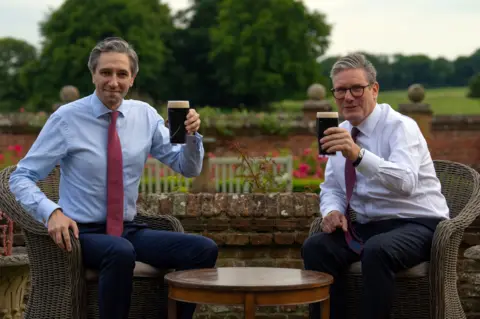 bbc 2  men successful  shirts and ties raising afloat  pint glasses of Guinness successful  the absorption  of the camera. They are sitting successful  wicker chairs with roses and trees successful  the background, and are connected  either broadside  of a circular  woody  table