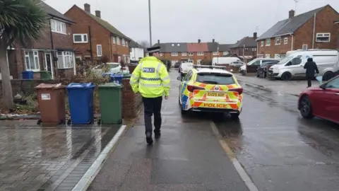 Essex Police A police car is parked on a residential street and partly on the pavement. The roads are wet with rain. One officer is seen from behind walking down the pavement, wearing a neon high vis coat, black trousers and a black police hat.