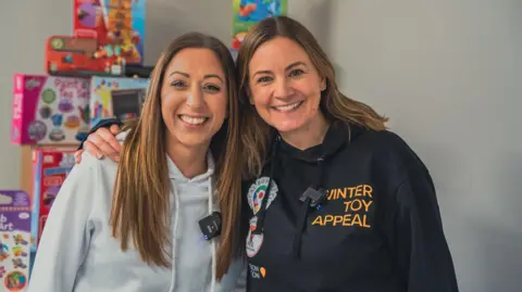 Winter Toy Appeal Vicky Bibiris (left) and Jenna Fansa (right) stand together smiling at the camera.
Vicky has long brown hair and wears a white sweater. Jenna has lighter, long hair and wears a black sweater, Toys are piled up in the background.