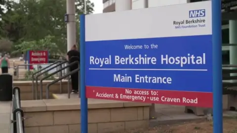 Large blue-and-white NHS sign outside the Royal Berkshire Hospital. The sign reads ‘Welcome to the Royal Berkshire Hospital – Main Entrance’ and includes a notice directing Accident and Emergency visitors to use Craven Road. Hospital buildings, railings and pedestrians are visible in the background.