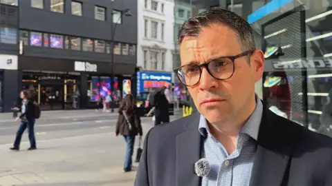A man with dark framed glasses in a dark blue suit and checked shirt standing on a high street, with shops and people in the background