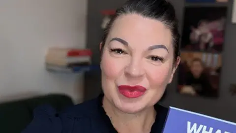 Kiki Deville smiles at the camera holding her book, which has a purple cover and is illustrated with a love-heart affixed with a sticking plaster