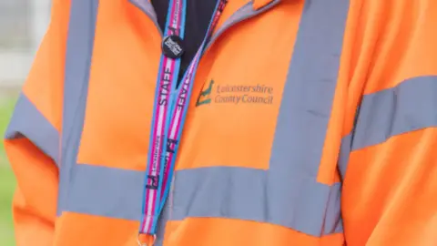 Leicestershire County Council An unidentifiable council staff member wearing a light blue, pink, and white lanyard over a high-vis orange coat