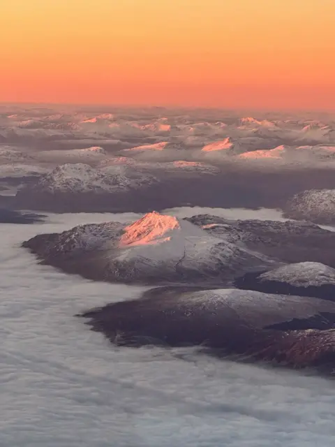 Alex Brand Image taken from a plane of snow covered hills peaking over the clouds as the sun sets