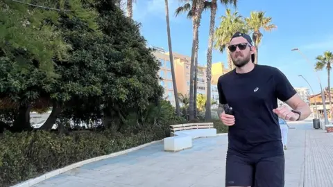 Brain Tumour Trust Alex with dark hair and beard wearing a black running strip and running past green trees in a city centre