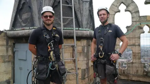 Joe Bilton/BBC Two men in black tops and shorts are wearing white helmets and harnesses. They are standing on the roof of a cathedral's central tower, next to a ladder which is leaning against a grey pinnacle.