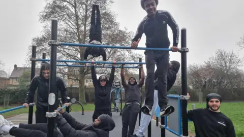 A group of people use the outdoor bar‑based fitness structure in a park. Some individuals are hanging from the bars, one is positioned upside down, and another is holding themselves above the bar. Others stand or lean on various parts of the structure. The scene takes place on a rubber‑surfaced exercise area with trees and grass in the background.