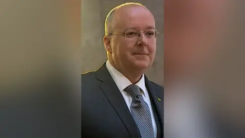 Former SNP chief executive Peter Murrell smiles as he looks off to the side. He is wearing glasses, a white shirt, dark grey blazer and a black and white tie.