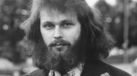 Getty Images A black and white photograph of Clark Olofsson in the 1970s. He is dressed in a flowery shirt with a dark suit. He has a bushy beard and is staring a the camera.
Olofsson